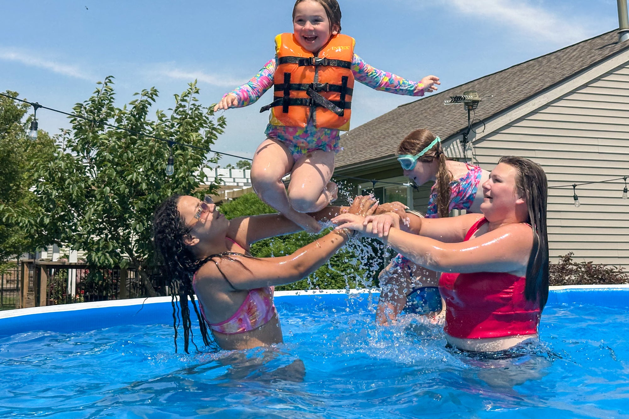 Family enjoying a Perfect Pool, with a woman and girl tossing a child wearing a life jacket into the water, demonstrating affordable above‑ground pool ownership and family fun