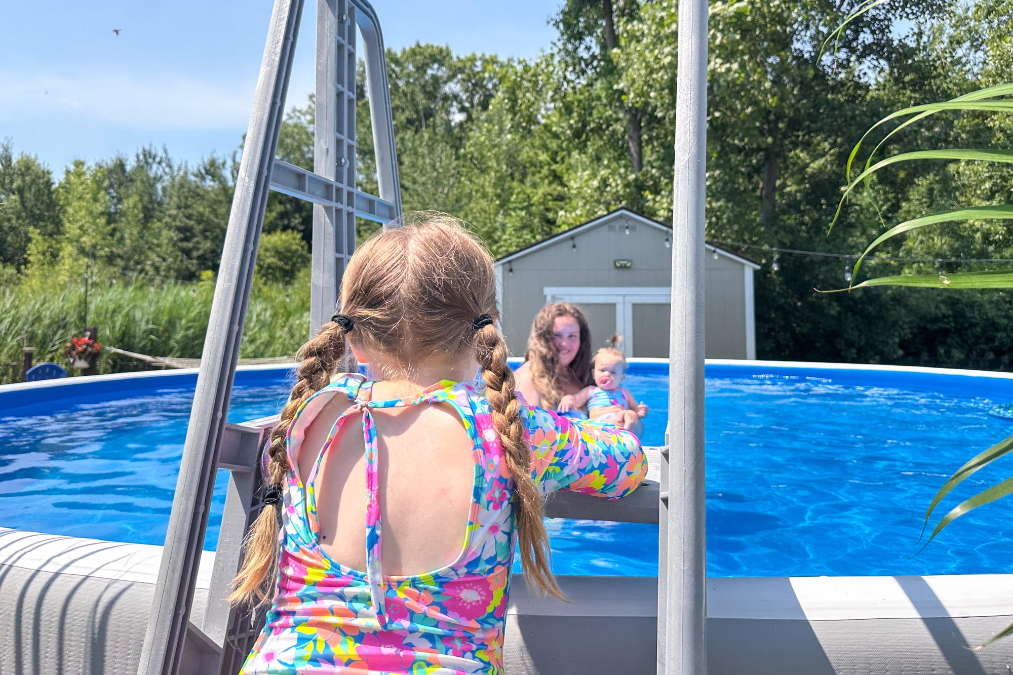 Young girl with braided pigtails climbing a ladder into a Perfect Pool while her mother holds a baby in the water, showing the pool’s family‑friendly design and sturdy ladder