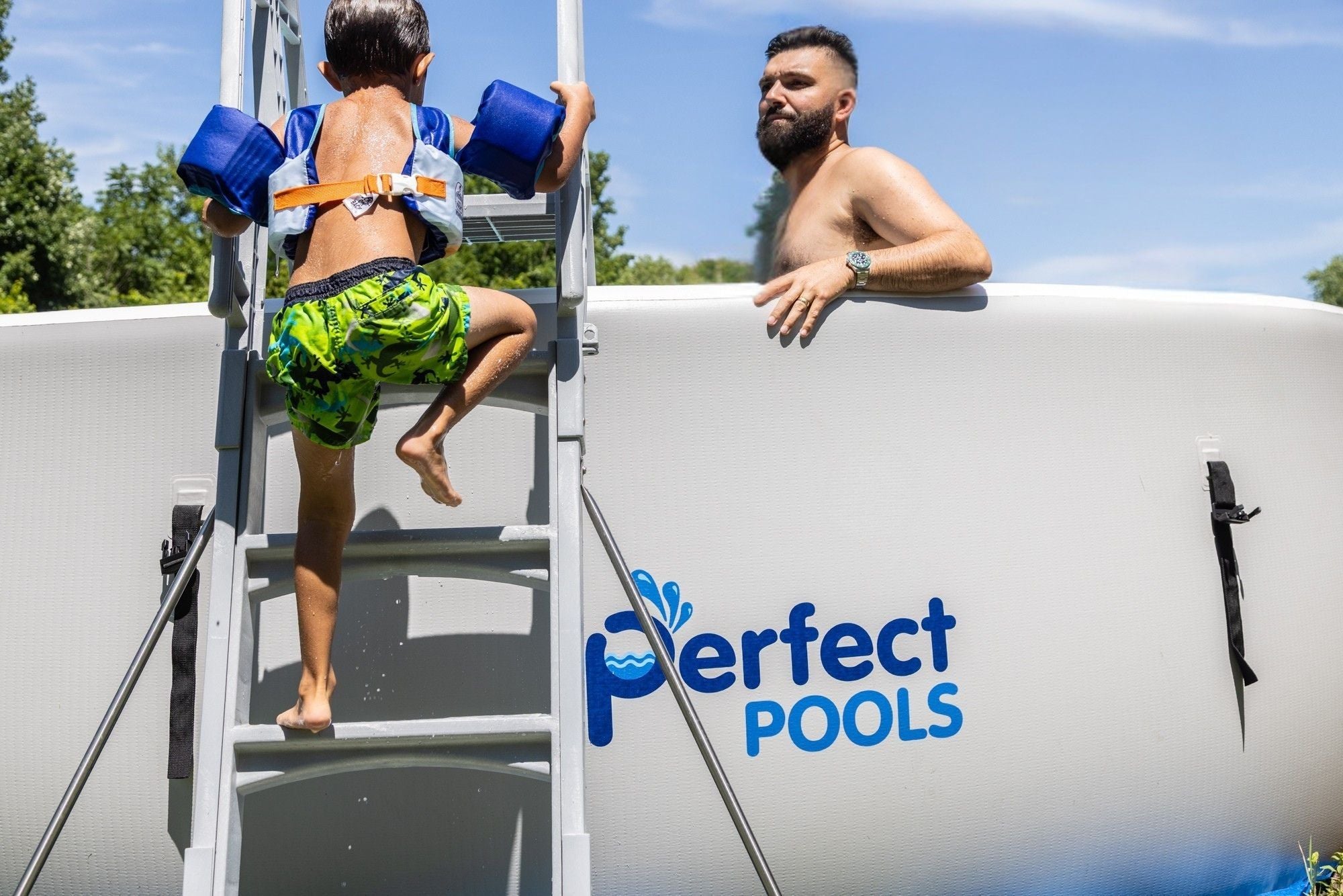 Young boy wearing blue flotation armbands climbing a ladder into a Perfect Pool as a bearded man supervises from inside the pool, with the Perfect Pools logo visible on the pool wall
