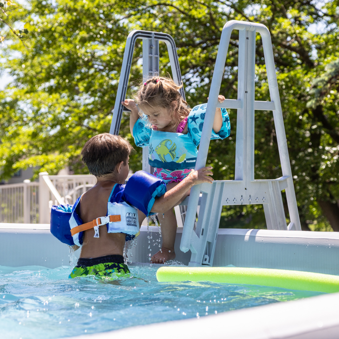 Boy in the water helping a younger girl in a flotation vest climb the Perfect Pool ladder, demonstrating family fun and safety