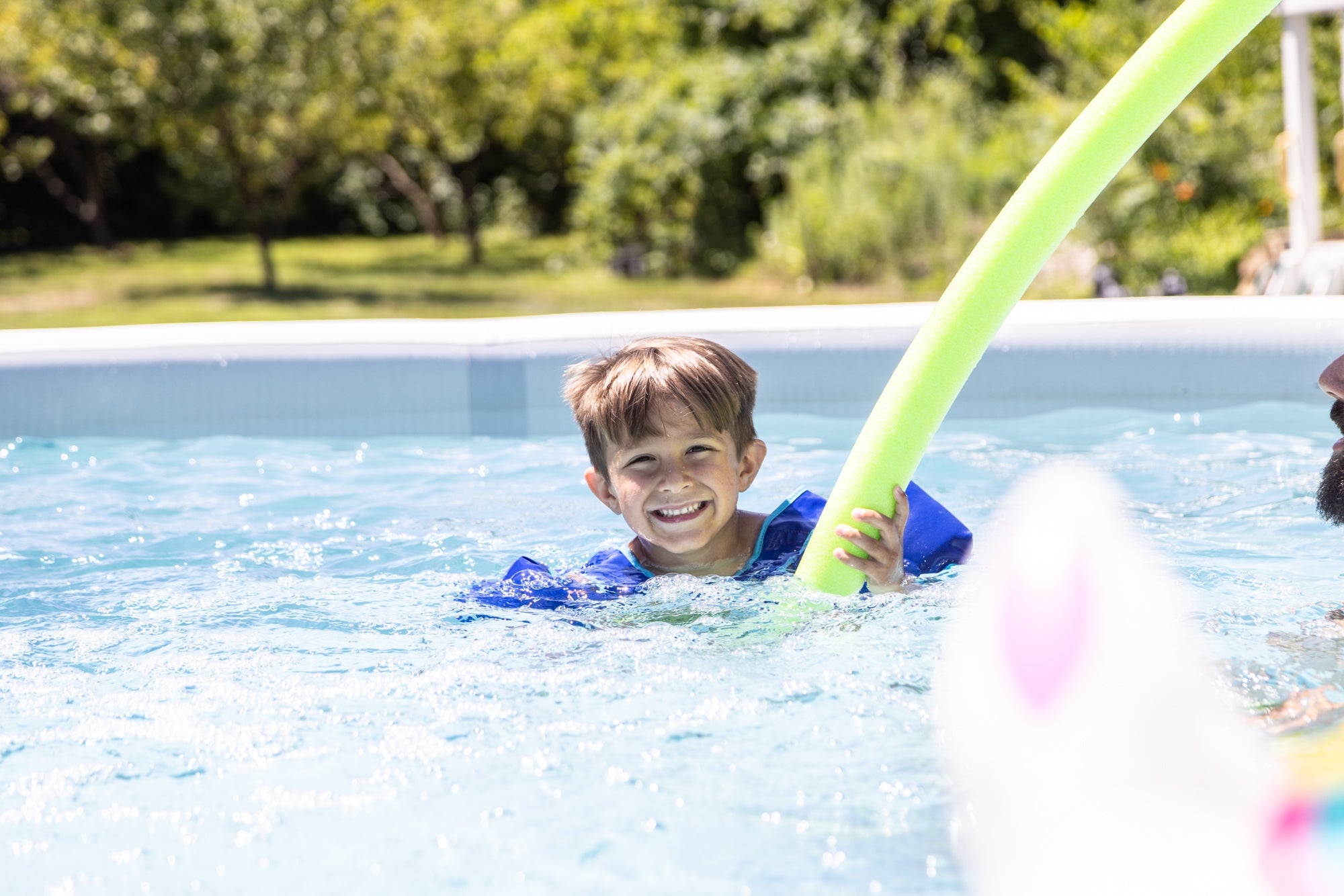 Smiling boy wearing a blue flotation vest and holding a green pool noodle as he plays in a Perfect Pool above‑ground pool