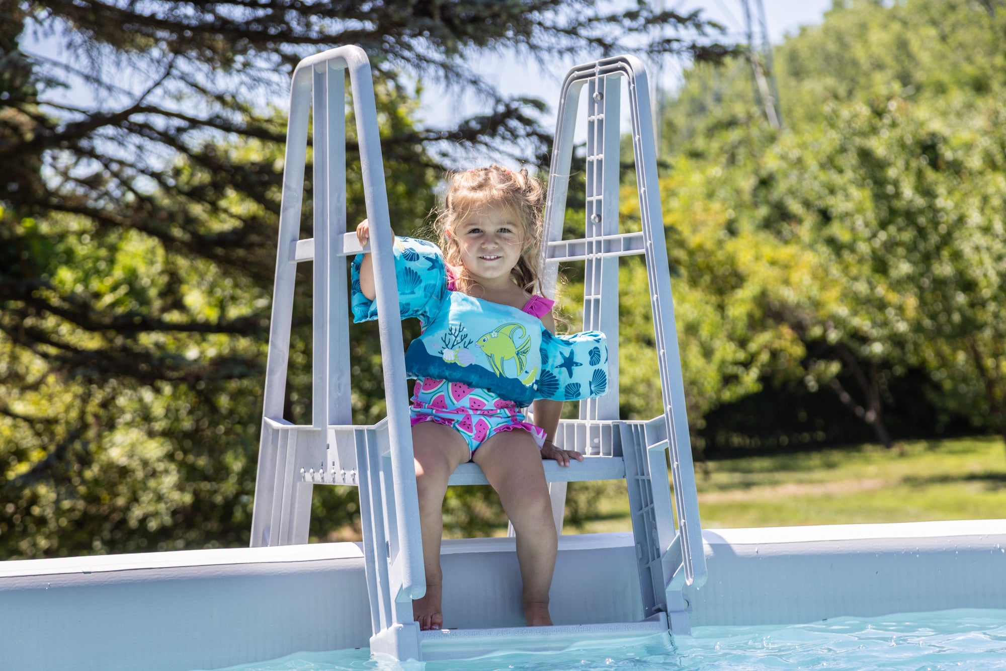 Young girl wearing colorful flotation sleeves sitting on a Perfect Pool ladder, getting ready to swim